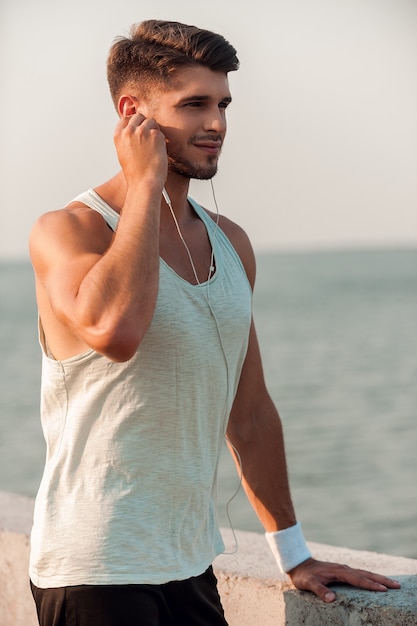 Buena música es todo lo que necesito para correr. Hombre musculoso joven confiado en auriculares apoyado en el parapeto y mirando a otro lado mientras está de pie al aire libre