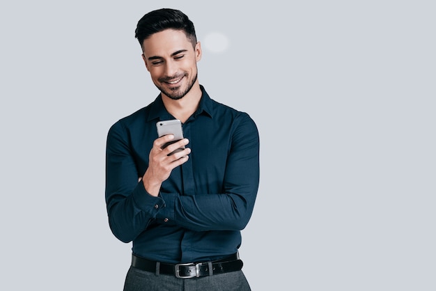 Buenas noticias de un amigo. Apuesto joven sosteniendo un teléfono inteligente y mirándolo con una sonrisa