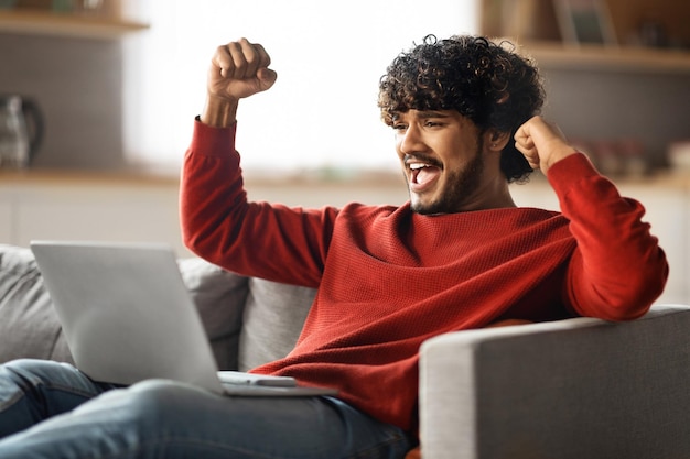 Buenas noticias feliz emocionado hombre indio celebrando el éxito con una computadora portátil en casa