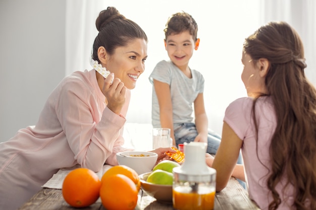 Buenas vitaminas. Atractiva madre joven de pelo oscuro sonriente sosteniendo vitaminas y hablando con sus hijos sobre la atención médica y el niño sentado en la mesa