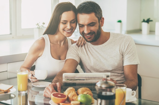 Buenos días comienza con buenas noticias. Hermosa joven pareja uniéndose y leyendo el periódico juntos mientras está sentado en la cocina y desayunando