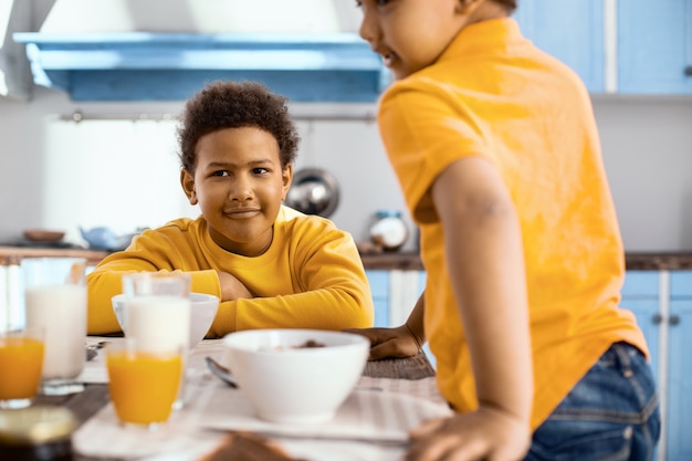 Buenos días. Encantador niño preadolescente sentado a la mesa y desayunando mientras su hermano menor se une a él en la mesa