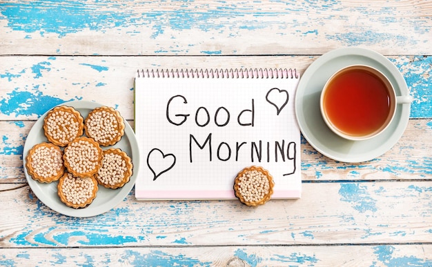 "Buenos días" está escrito en el bloc de notas con una taza de té y galletas en la mesa. Vista superior.