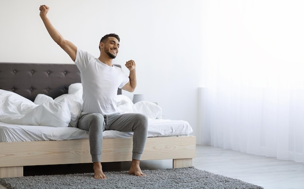 Buenos días, feliz árabe sentado en la cama y estirándose después de dormir.