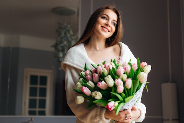 ¡Buenos días! Mujer joven atractiva con ramo de tulipanes blancos y rosas pasa tiempo en casa.