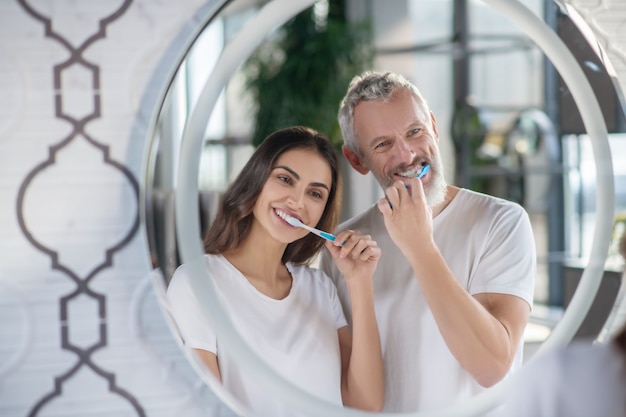 Buenos días. Mujer sonriente y hombre de pie uno al lado del otro reflejado en el espejo cepillarse los dientes