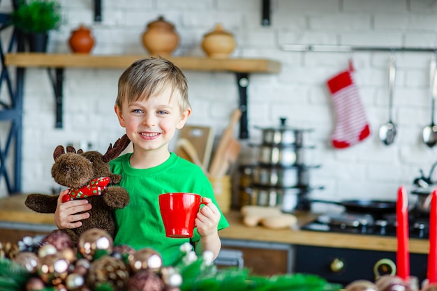 Buenos días. Un niño bebe té en la mesa de la cocina y abraza a un alce de peluche. Un tiempo de milagros y cumplimiento de deseos.