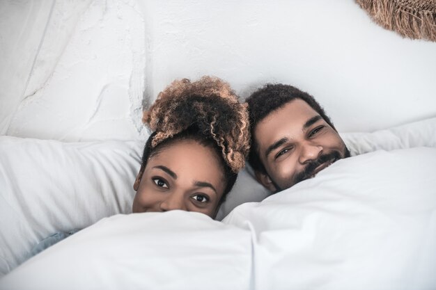 Buenos dias. Sonriente hombre y mujer felices de piel oscura que yacen bajo una manta blanca, solo las caras son visibles