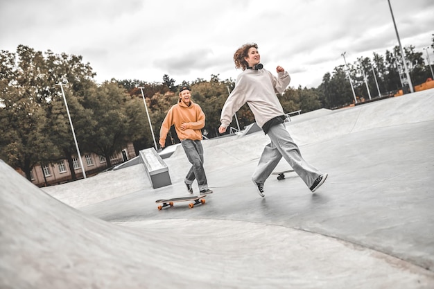 Buenos momentos. Chico pelirrojo sonriente con gorra corriendo detrás de una chica alegre con collares cerca de patinetas de pie en el skatepark en la tarde