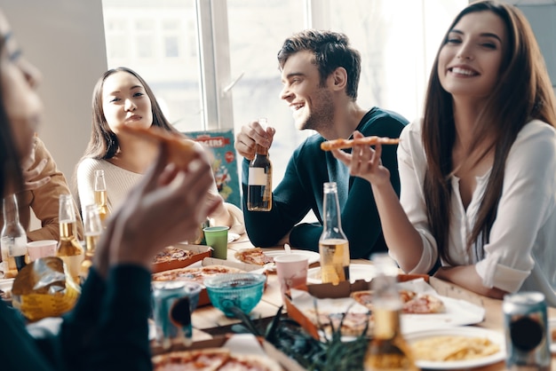 Buenos tiempos. Grupo de jóvenes en ropa casual comiendo pizza y sonriendo mientras tienen una cena en el interior