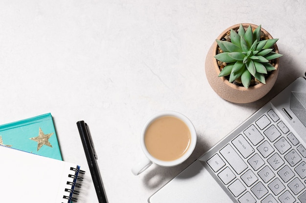 Büro-Desktop aus weißem Marmor mit Laptop, Notizbuch, Tasse Kaffee und saftiger Blume in einer Kanne