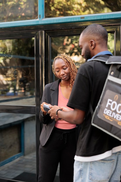 Büroangestellte bezahlen für die Lieferung von Lebensmitteln mit POS-Terminal, kontaktlose Zahlung per Kreditkarte. Afroamerikanischer Kurier mit Rucksack, der dem Kunden in der Nähe des Firmengebäudes eine Mahlzeit im Restaurant gibt