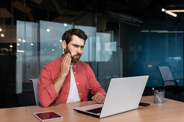 Büroangestellter hat Zahnschmerzen, ein kranker Geschäftsmann arbeitet im Büro mit einem Laptop, den ein Mann hat