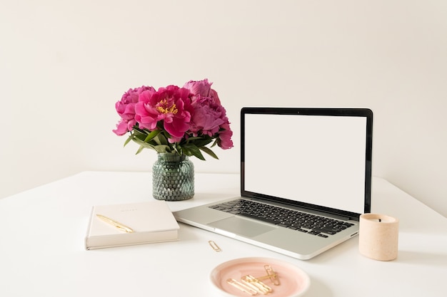 Büroarbeitsplatz Tischarbeitsplatz mit leerem Kopierraum Modell Laptop-Bildschirm. Modernes stilvolles Hauptinnendesign mit Blumenstrauß der rosa Pfingstrosen.
