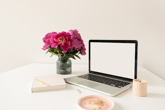 Büroarbeitsplatz Tischarbeitsplatz mit leerem Kopierraum Modell Laptop-Bildschirm. Modernes stilvolles Hauptinnendesign mit Blumenstrauß der rosa Pfingstrosen.