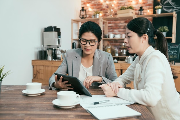 Bürodamen Geschäftskooperation treffen Teamwork-Konzept. Zwei Geschäftsfrauen unterhalten sich am Tisch im Café-Shop. Mädchen im Anzug zeigt Kollegeninformationen auf dem Tablet-Bildschirm, der in der Kaffeebar arbeitet