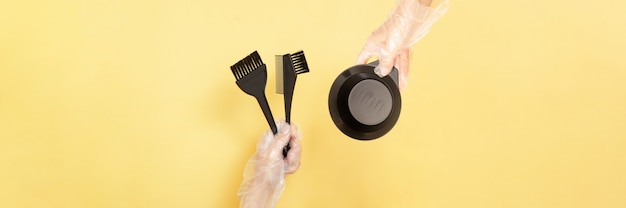 Bürsten und Schüssel zum Färben von Haaren zu Hause oder im Salon in den Händen einer Frau mit Handschuhen auf gelbem Hintergrund