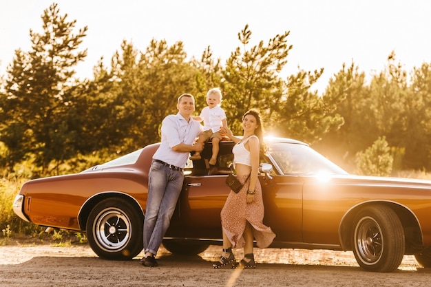 Buick Rivierin Retro-Stil. Einzigartiges Auto. Netter blonder Junge, der auf dem Dach des Autos sitzt. Eltern umarmen ihren Sonnenuntergang.