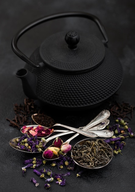 Bule de chá chinês com xícara e colheres com vários chás em fundo preto Botões de rosas flores de malva azulchá verde e preto a granel