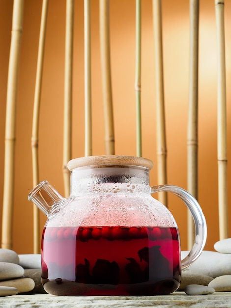 Foto bule de chá de vidro com karkade de chá quente de frutas vermelhas, close-up. chá no fundo de um bosque de bambu e pedras. conceito de festa do chá chinês tradicional. dia internacional do chá, plano de fundo.