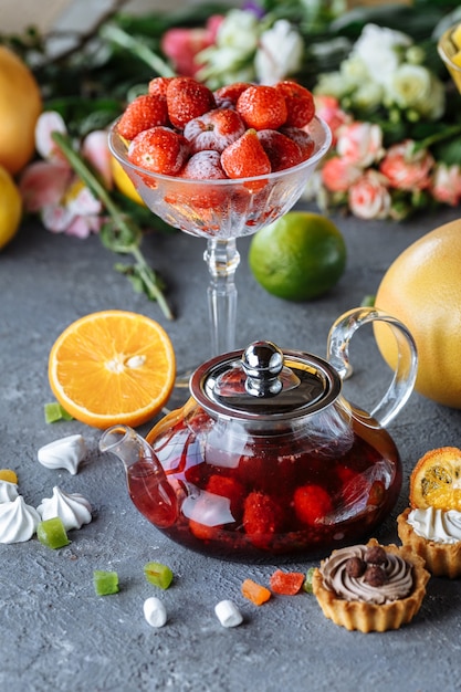 Bule de vidro com chá de frutas framboesa e hortelã em um fundo azul com frutas e decorações.