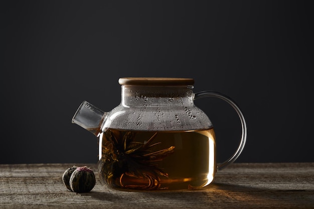Bule transparente com chá florescente e bolas de chá em mesa de madeira isolada em preto