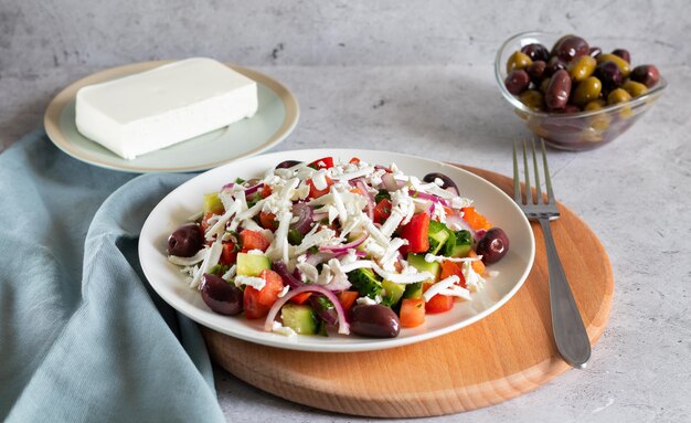 Bulgarischer Schopska-Salat mit Gemüse und Käse auf hellem Betongrund und Holzbrett mit grauen Textilien und Gabel Horizontale Seitenansicht