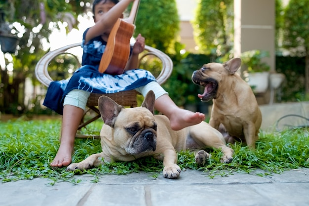 Bulldog francés acostado junto a una niña pequeña que toca el ukelele en el jardín