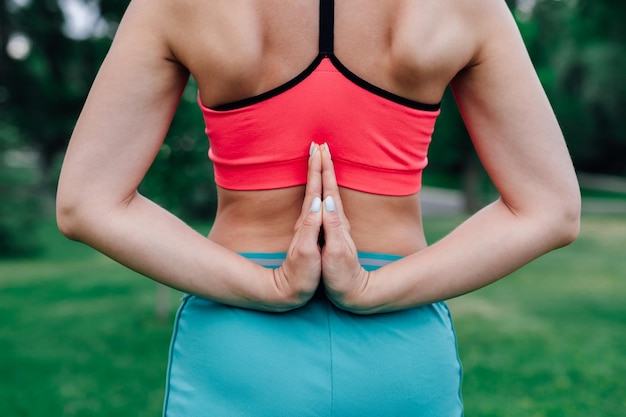 Übungen für die richtige Körperhaltung Körper einer jungen Frau in rosa Oberteil und blauer Sporthose mit hinter dem Rücken gefalteten Handflächen in Pose von Paschima Namaskarasanart Asana Rückansicht