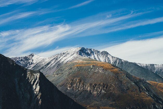 Bunte Alpenlandschaft mit großem Berg in Herbstfarben mit Schnee auf dem Gipfel bei Sonnenschein unter Zirruswolken am blauen Himmel. Malerische Herbstlandschaft mit sonnenbeschienenen Felsen und schneebedeckten Berggipfeln