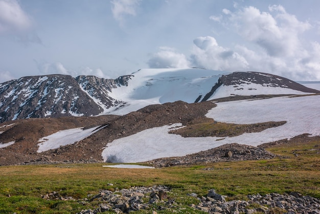 Bunte grüne Landschaft mit Wolkenschatten auf sonnenbeschienenem, schneebedecktem Berggipfel unter bewölktem Himmel bei wechselhaftem Wetter. Fantastische sonnige Landschaft mit See in Graswiese vor hoher schneebedeckter Bergkette