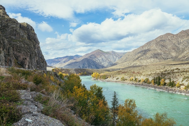 Bunte Herbstlandschaft mit goldenen Blättern auf Bäumen entlang des breiten türkisfarbenen Bergflusses bei Sonnenschein. Helle Alpenlandschaft mit großem Bergfluss und gelben Bäumen in goldenen Herbstfarben im Herbst