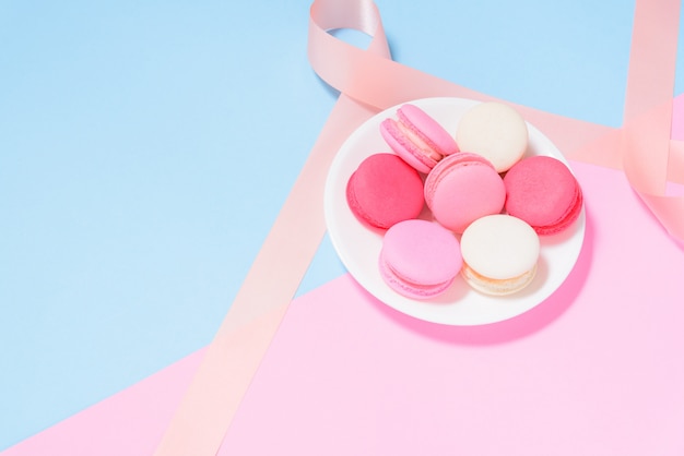 Bunte Makronen oder Macaron auf weißer Platte mit rosa Band auf rosa und blauem Pastellhintergrund