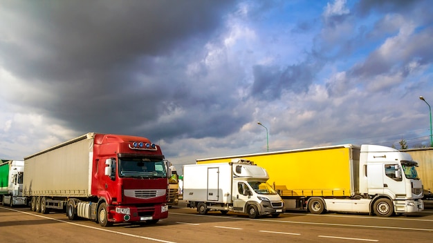 Bunte moderne große Sattelzugmaschinen und Anhänger verschiedener Marken und Modelle stehen in Reihe auf dem flachen Parkplatz der Lkw-Haltestelle bei Sonnenschein