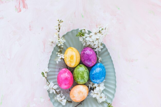 Bunte Ostereier auf dem Teller und weiße Kirschblüten im Frühling auf rosa Hintergrund Osterfeierkonzept Draufsicht flach liegender Kopierraum