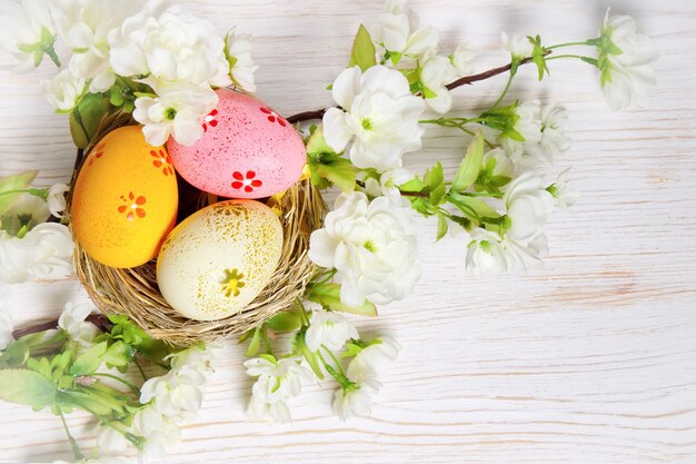 Bunte Ostereier im Strohnest und Zweig mit Blumen auf weißem Holztisch. Draufsicht, flach liegen.