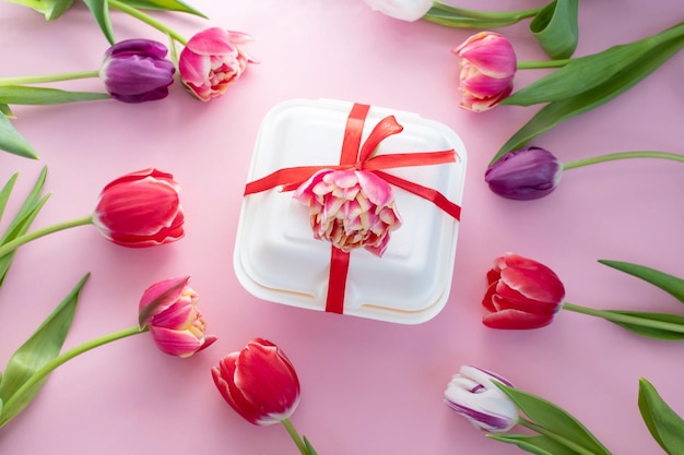 Bunte rote doppelte Tulpen liegen um eine weiße Dessertgeschenkbox mit einem roten Band Ansicht von oben