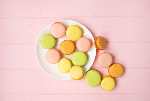 Bunter französischer oder italienischer Macarons-Stapel auf weißem Teller, der auf rosa Holztisch mit Kopienraum für Hintergrund gesetzt wird. Dessert zum Servieren mit Nachmittagstee oder Kaffeepause.