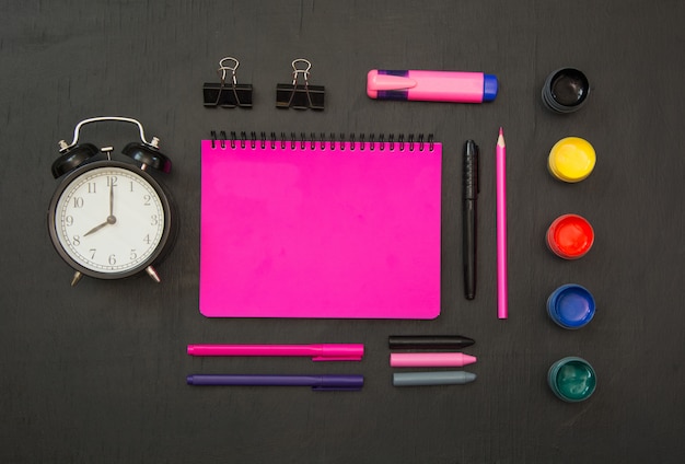 Bunter Schulbedarf, purpurrotes Schreibheft und Wecker auf schwarzer Tafel.