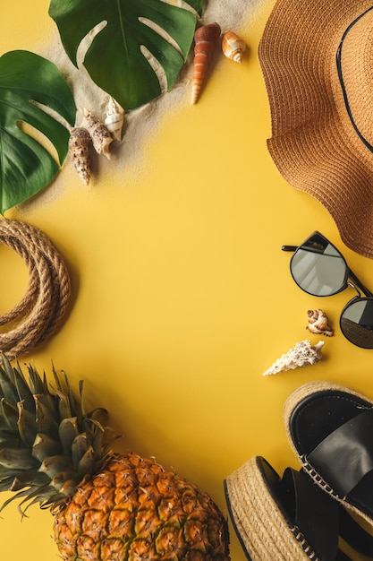 Buntes Sommermode-Outfit flach lag Strohhut Korbschuhe Ananas über gelbem Hintergrund Draufsicht breite Komposition Sommermode-Urlaubskonzept