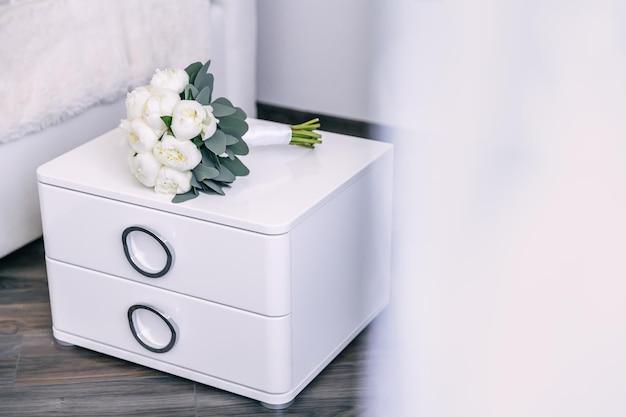 Buquê de casamento deitado em uma cômoda branca enquanto se prepara para a celebração decorativa flores de peônia branca e buquê de folhas