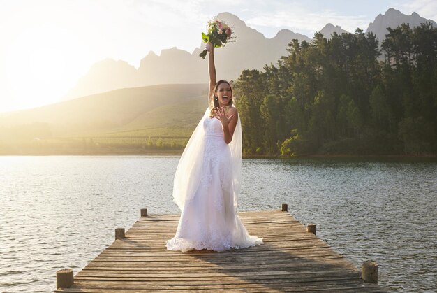 Buquê de casamento e celebração com uma noiva em um píer sobre um lago na natureza após uma cerimônia de casamento sorriso feliz e flores com uma mulher comemorando o casamento na tradição do matrimônio