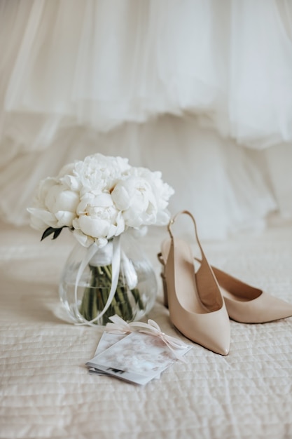 Buquê de flores de peônias em um vaso fica na cama dos noivos com convites e sapatos no espaço dos vestidos de noiva
