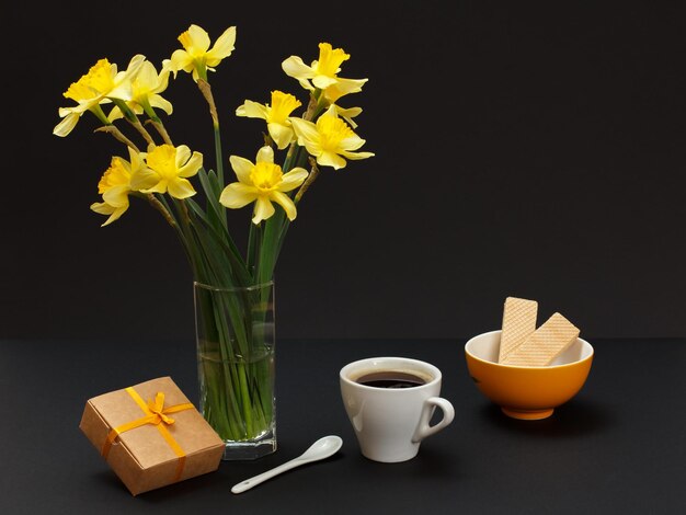 Buquê de narcisos amarelos em vaso de vidro uma caixa de presente uma xícara de café e um prato com waffles