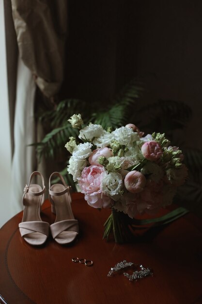 Buquê de noiva de rosas brancas e peônias com sapatos de noiva e anéis de casamento na mesa antes da cerimônia de casamento