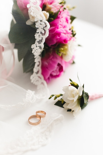 Buquê de peônias rosa linda e anéis de casamento mentem sobre uma mesa branca.