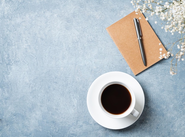 Buquê de primavera com flores brancas de gypsophila sobre uma xícara de café preto e bloco de notas de artesanato em uma mesa azul Conceito de 8 de março feliz dia da mulher em casa e local de trabalho no escritório Vista superior e espaço para cópia