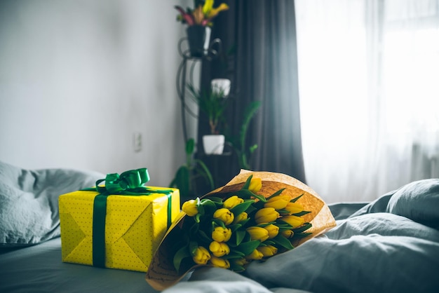 Buquê de tulipas amarelas com caixa de presente amarela na cama com lençóis cinza. a luz do sol da manhã brilha pela janela