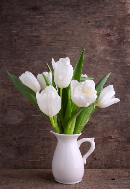 Buquê de tulipas brancas em um jarro branco sobre um fundo de madeira velho.