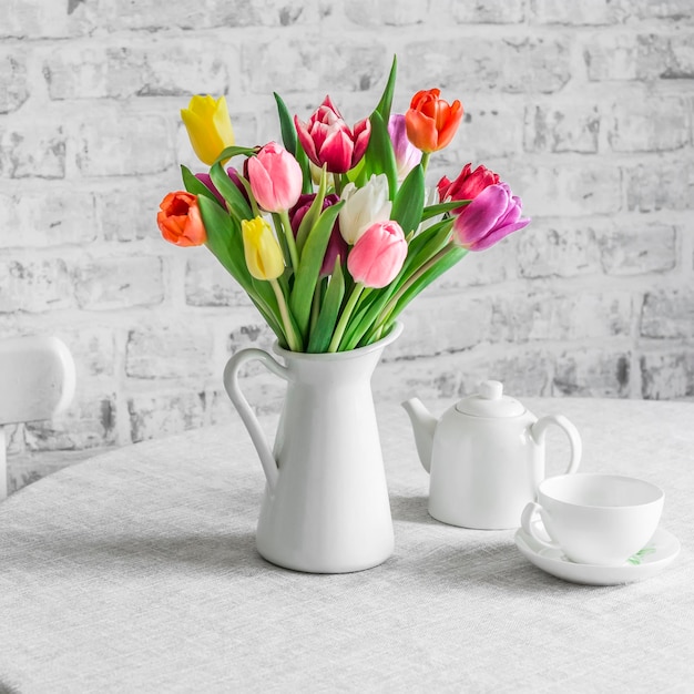 Buquê de tulipas coloridas, bule branco de cerâmica e xícara de chá na mesa na cozinha brilhante Conceito de casa aconchegante